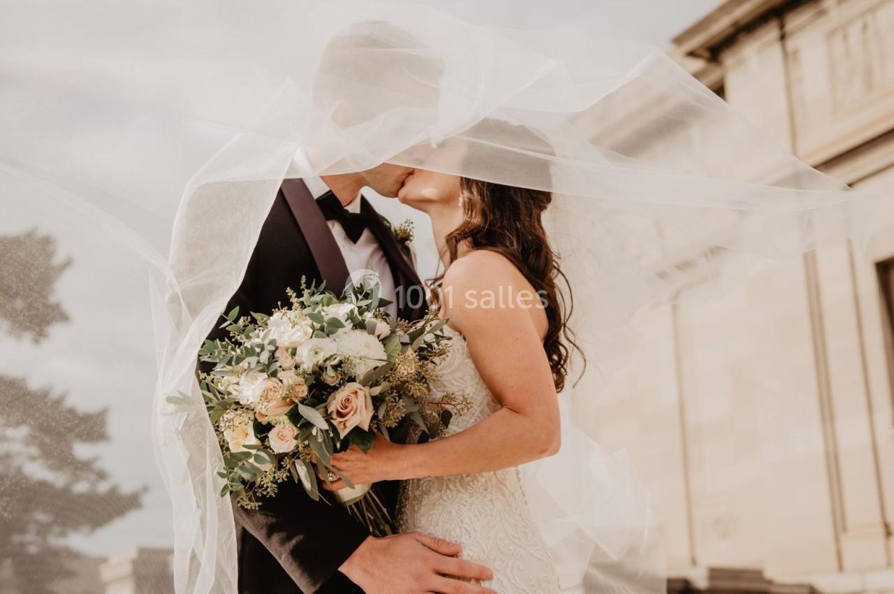 Un couple en tenue de mariage s'embrasse sous un voile, tenant un bouquet de fleurs, devant un bâtiment ancien.