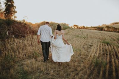 Un couple marche dans un champ au coucher du soleil, la femme portant une robe blanche et tenant légèrement son ourlet.