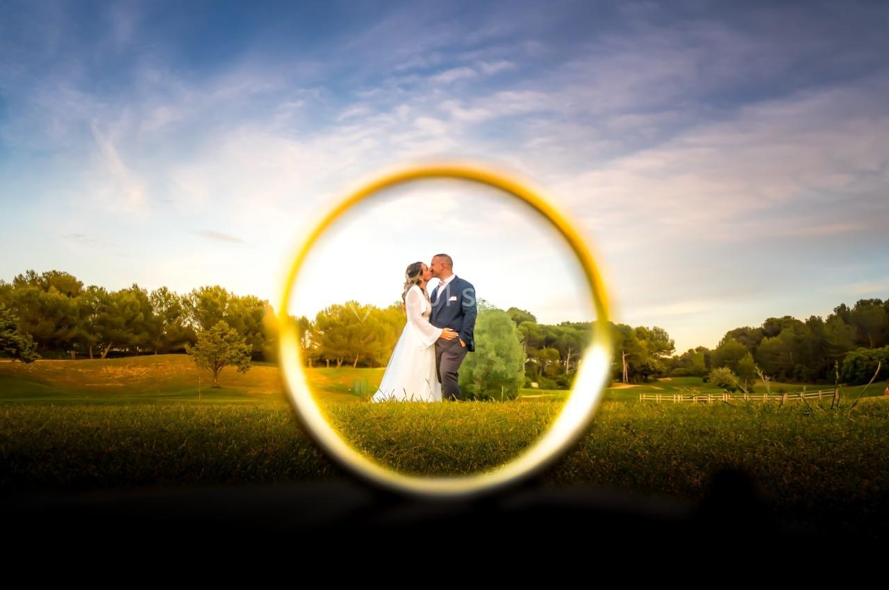Un couple en tenue de mariage se tient dans un parc, vu à travers une bague dorée en premier plan.