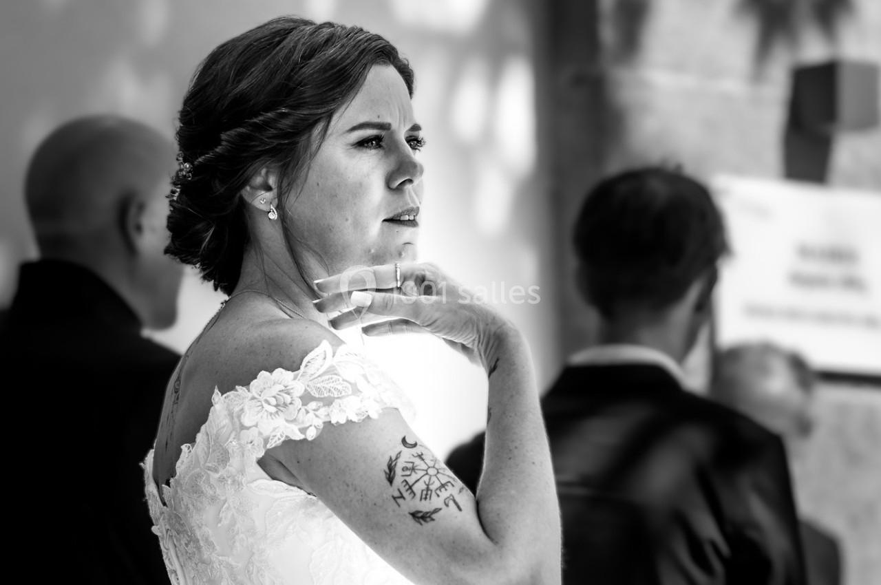 Femme en robe de mariée blanche, vue de profil, entourée de personnes en arrière-plan flou.
