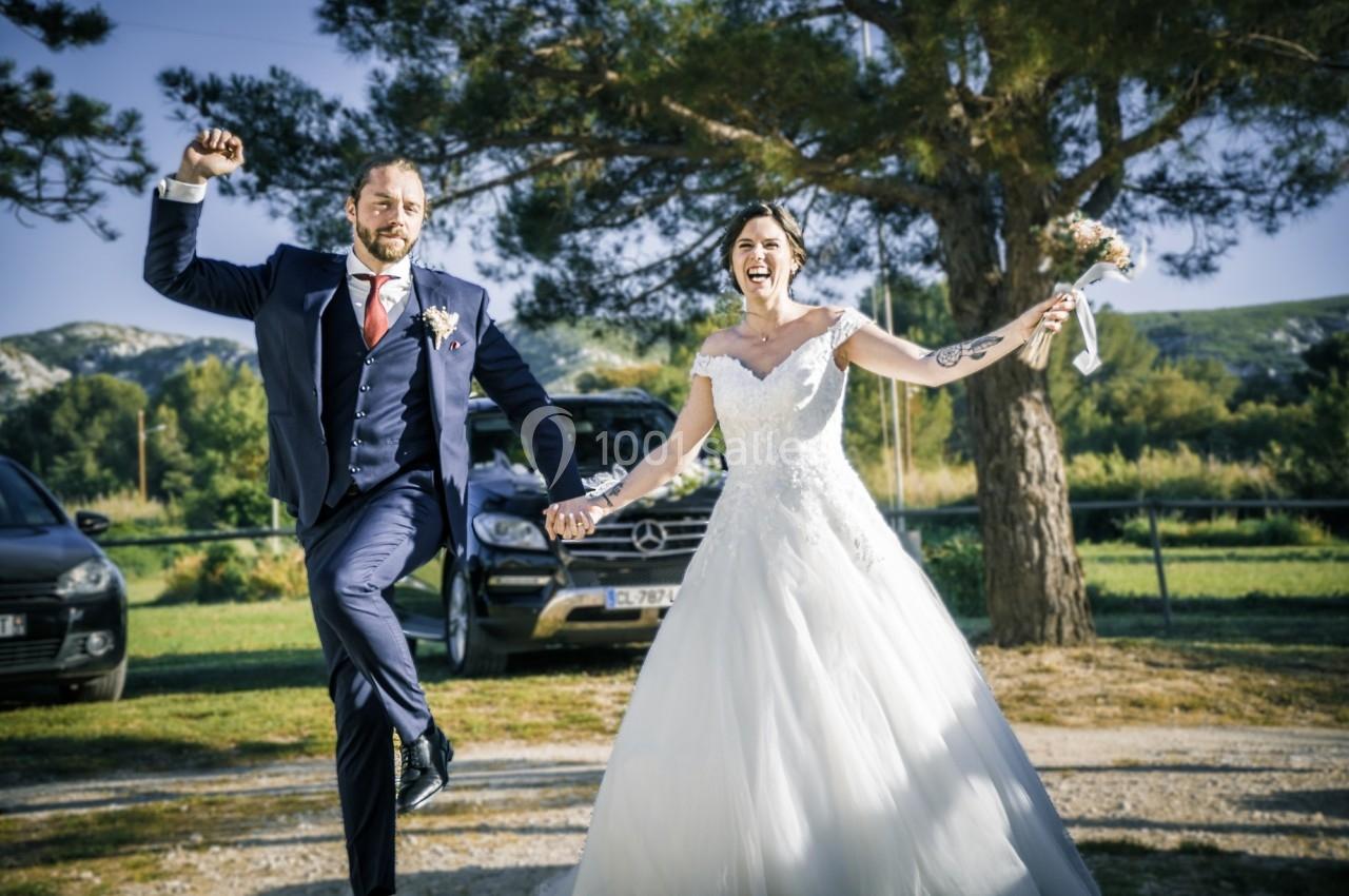 Un couple de mariés souriants pose joyeusement en extérieur, devant des arbres et des voitures.