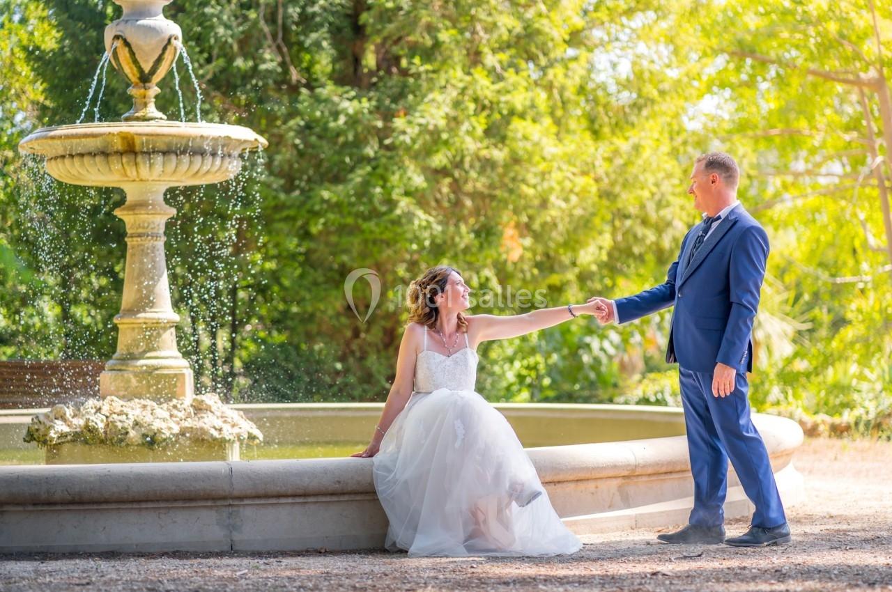 Une mariée assise au bord d'une fontaine tend la main à un homme en costume dans un jardin ensoleillé.