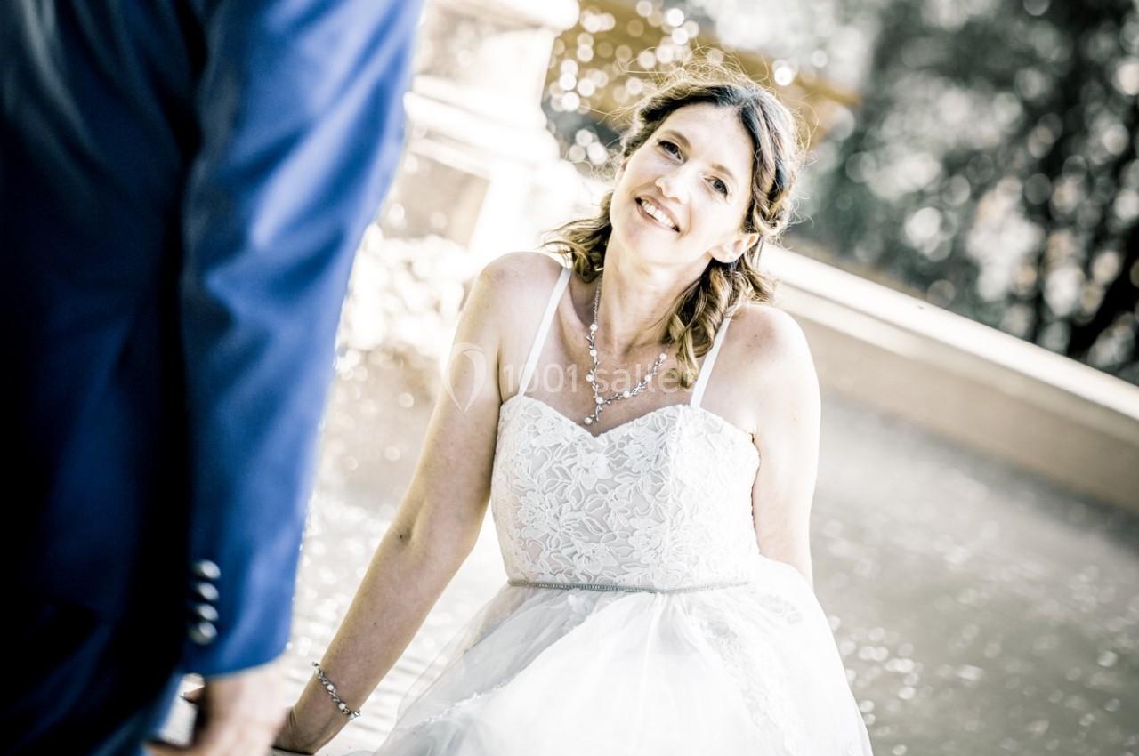 Femme en robe blanche souriante, assise près d'une fontaine, avec un homme en costume visible de dos.