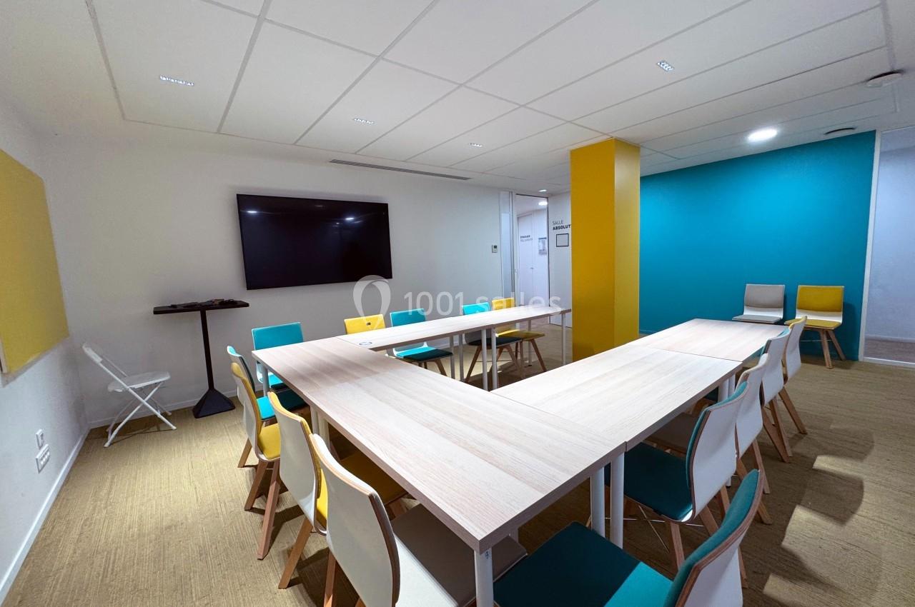 Salle de réunion lumineuse avec tables en forme de U, chaises colorées, écran mural et murs blancs et bleus.