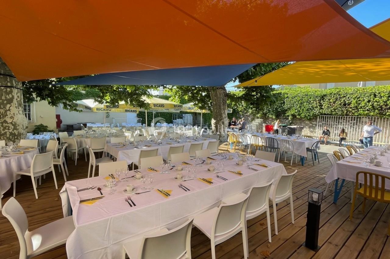 Tables dressées avec nappes blanches sous des voiles d'ombrage colorées, dans un espace extérieur entouré de verdure.