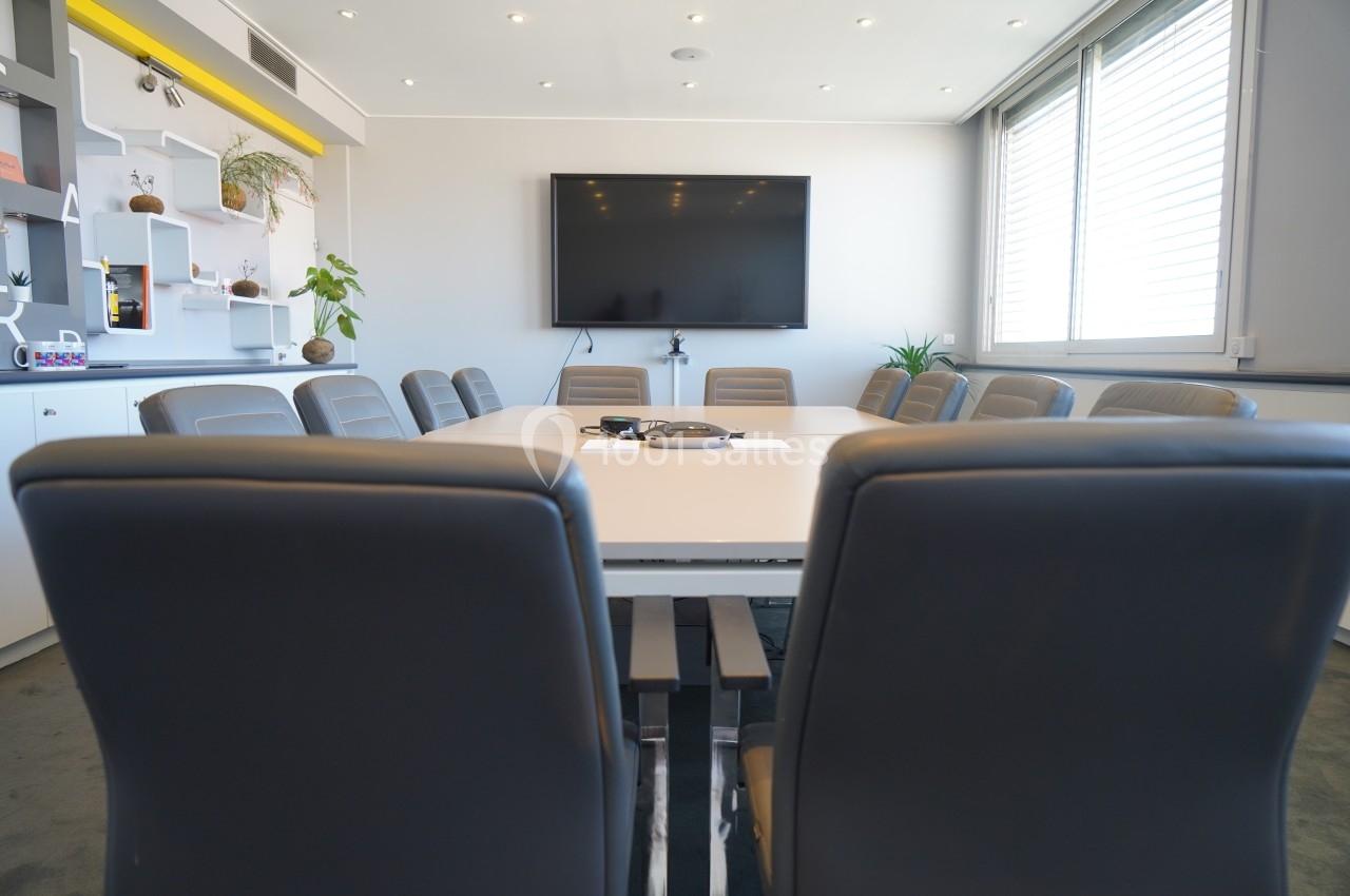 Salle de réunion moderne avec table blanche, chaises grises, écran mural et étagères décorées en arrière-plan.