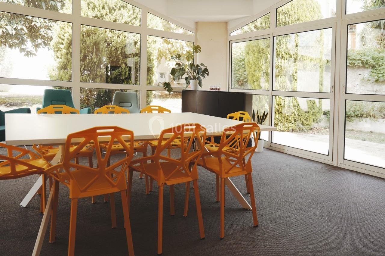 Salle de réunion lumineuse avec table blanche, chaises orange et grandes baies vitrées donnant sur un jardin.