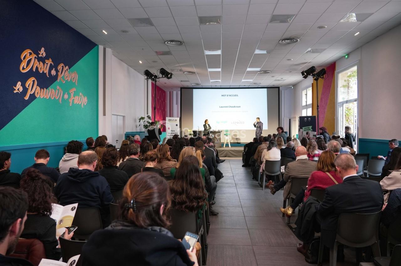 Salle de conférence avec un public attentif, un intervenant sur scène et une présentation projetée à l'écran.
