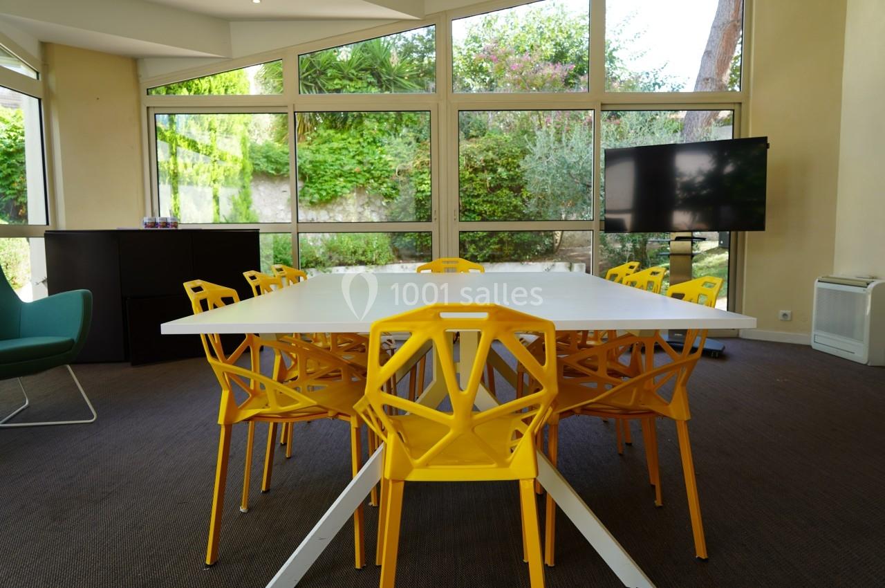 Salle de réunion lumineuse avec table blanche, chaises jaunes au design moderne et écran noir devant des baies vitrées.