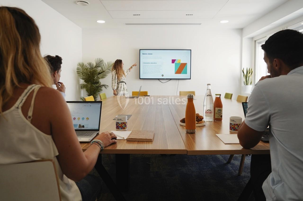 Une femme présente un contenu sur un écran dans une salle de réunion avec plusieurs personnes autour d'une table.