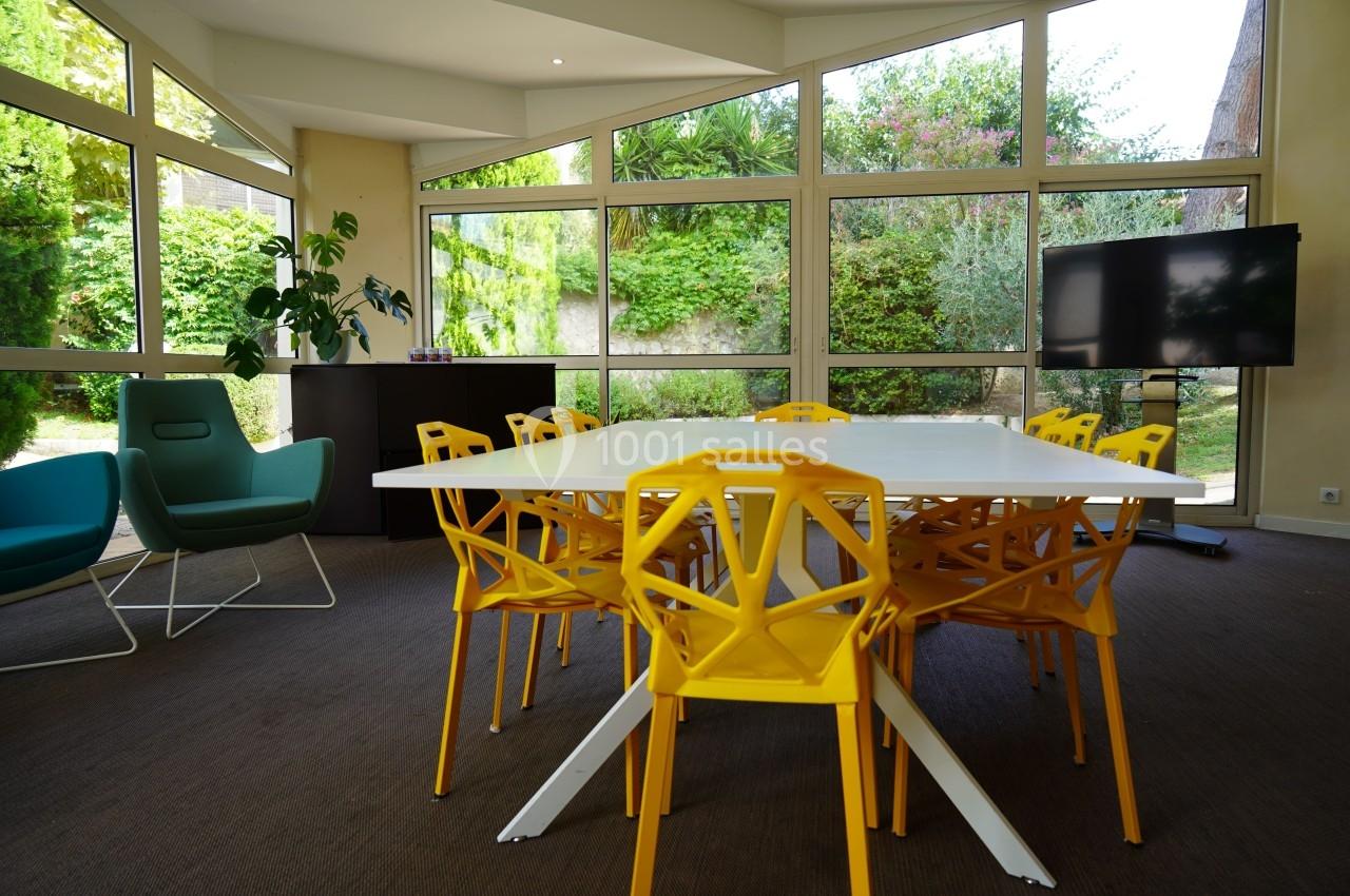 Salle lumineuse avec table blanche entourée de chaises jaunes, fauteuils et écran, donnant sur un jardin verdoyant.