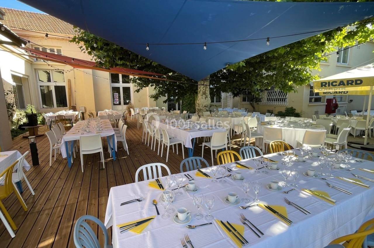 Terrasse en bois aménagée avec des tables dressées sous des voiles d'ombrage bleues et des arbres.