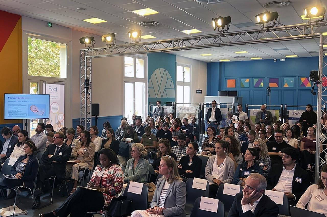 Salle de conférence remplie de participants assis, écoutant une présentation avec écran et affiches en arrière-plan.