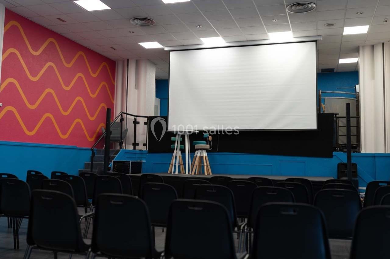 Salle de conférence avec des rangées de chaises noires, un écran de projection et deux tabourets sur scène.