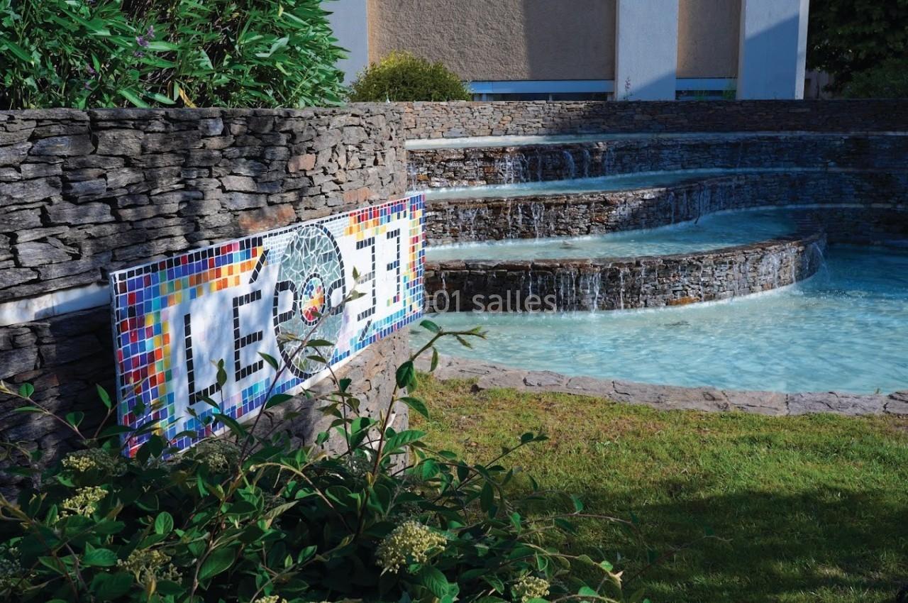 Mur en pierre avec une mosaïque colorée près d'une fontaine à plusieurs niveaux entourée de végétation.