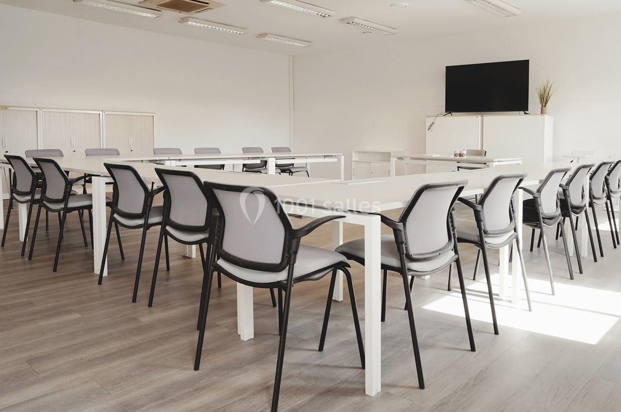 Salle de réunion lumineuse avec des tables blanches disposées en U, des chaises grises et un écran mural.