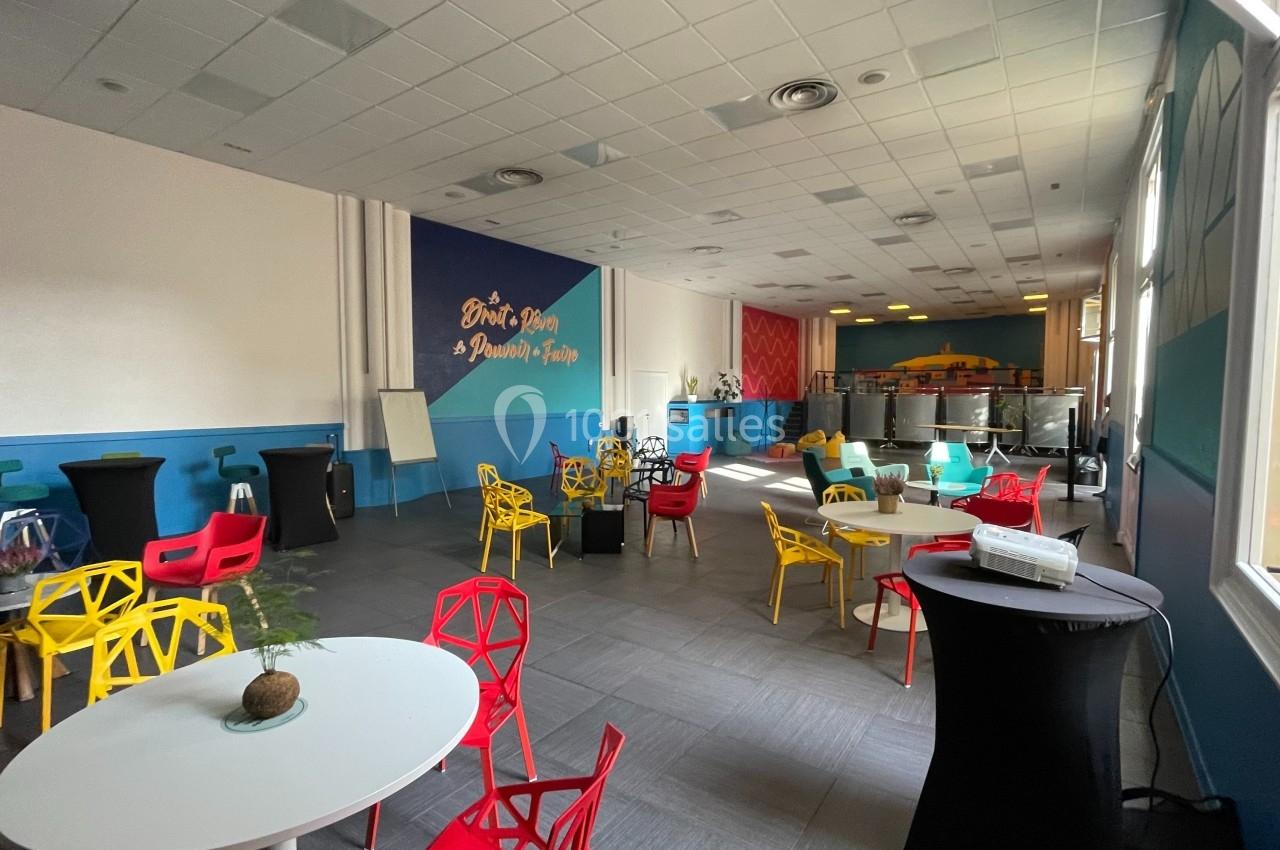 Salle lumineuse avec tables rondes blanches, chaises colorées et décorations modernes, vue sur un espace de restauration.