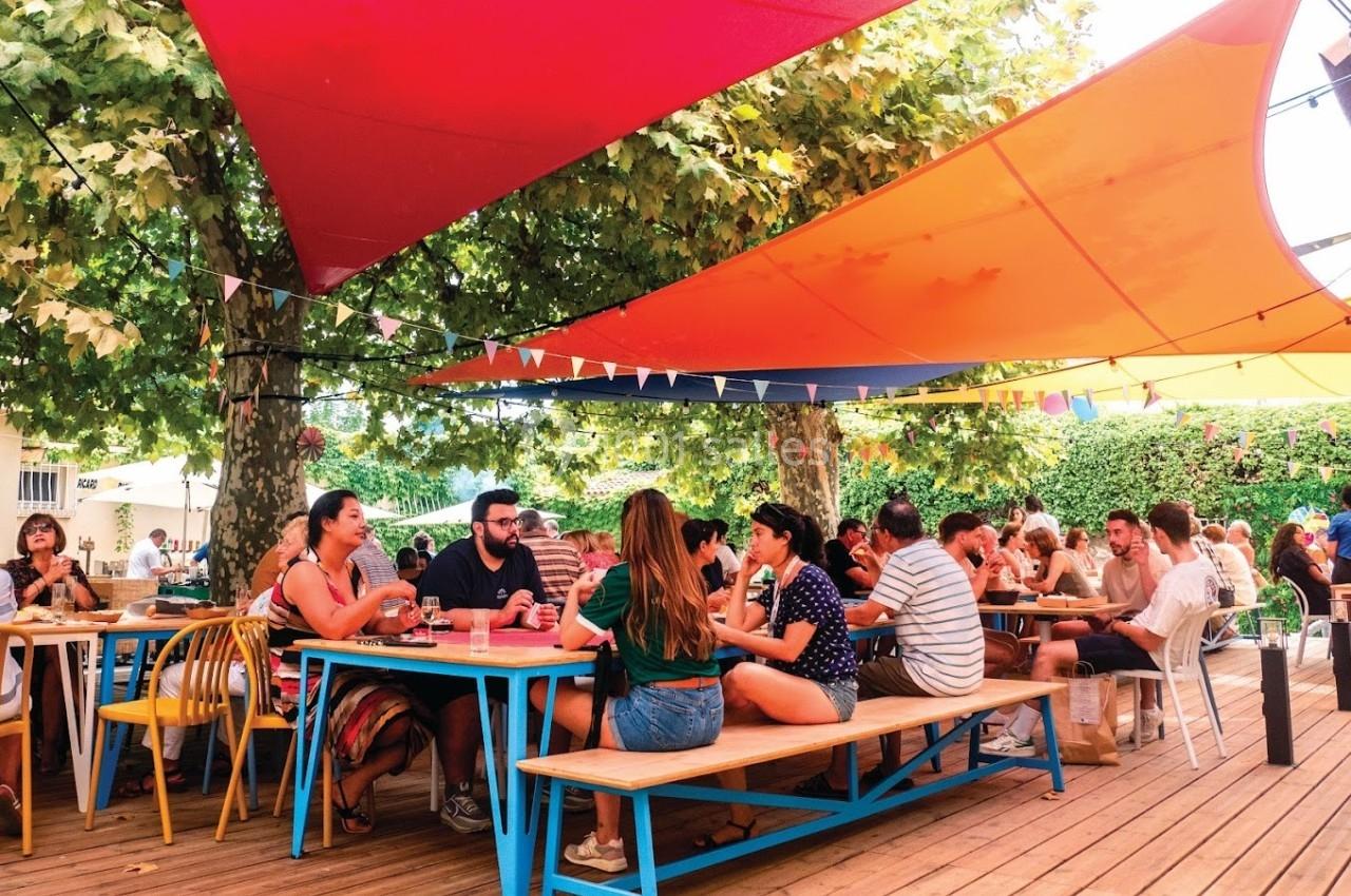 Des personnes discutent et partagent un repas à des tables en bois sous des voiles d'ombrage colorées en extérieur.
