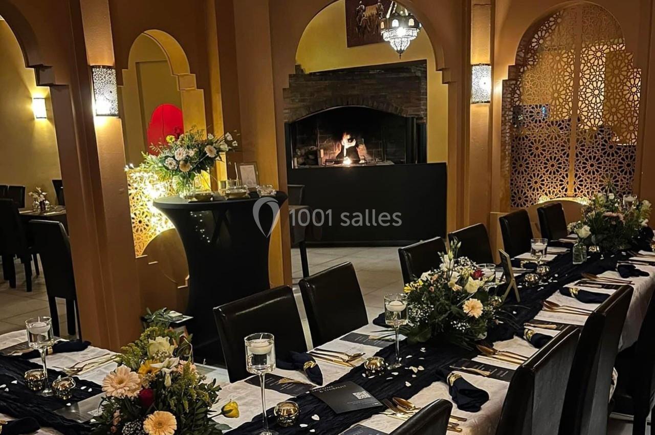 Salle de réception élégante avec tables dressées, décor floral, lumière tamisée et cheminée allumée en arrière-plan.