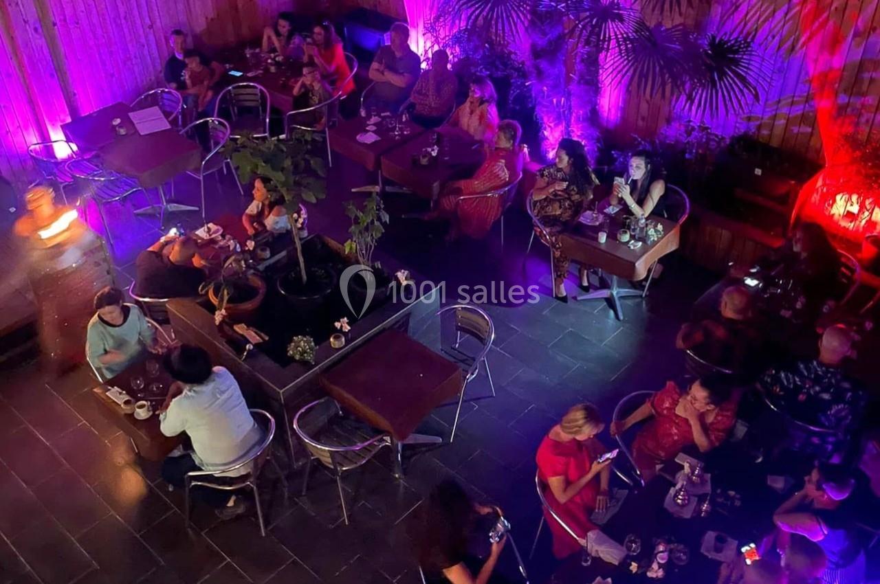 Vue plongeante d'une terrasse intérieure animée avec des tables occupées, éclairée par des lumières colorées.