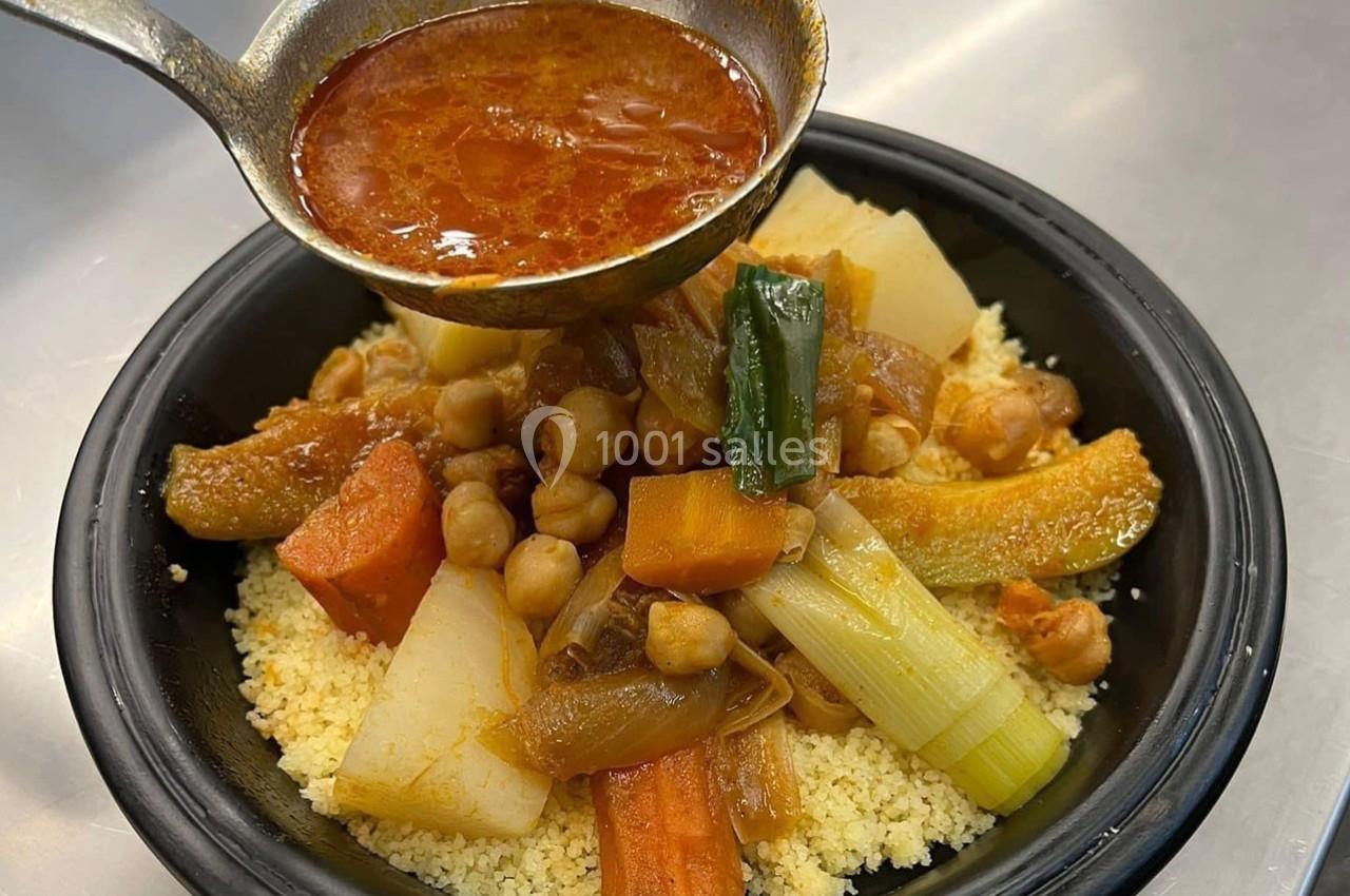 Un plat de couscous garni de légumes et pois chiches, avec une louche versant de la sauce rouge.