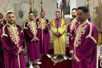 Un groupe d'hommes en tenues traditionnelles pose devant une décoration orientale et une affiche promotionnelle.