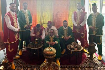 Un groupe d'hommes en tenues traditionnelles pose devant une décoration orientale et une affiche promotionnelle.