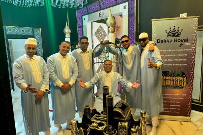 Un groupe d'hommes en tenues traditionnelles pose devant une décoration orientale et une affiche promotionnelle.