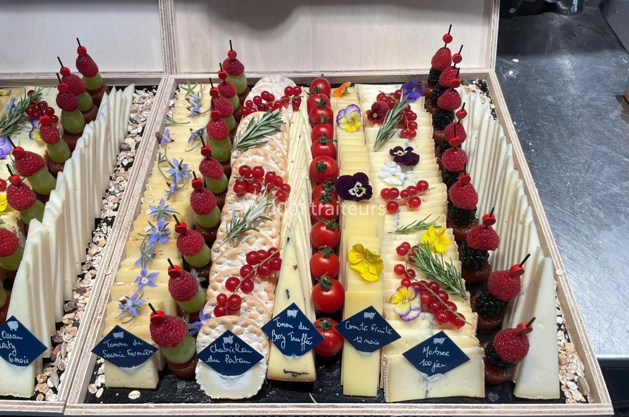 Plateau de fromages variés décorés de fruits, fleurs comestibles et herbes aromatiques, présenté dans un coffret en bois.