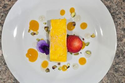 Dessert rectangulaire jaune avec fraise, pistaches, fleurs comestibles et sauces colorées sur une assiette blanche.