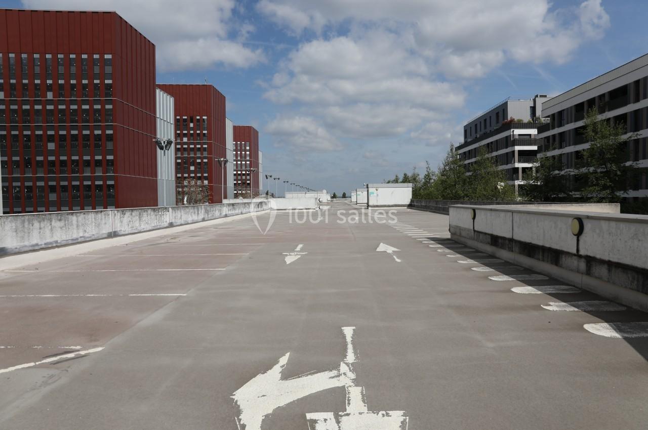Vue d'un parking vide en extérieur, entouré de bâtiments modernes sous un ciel partiellement nuageux.