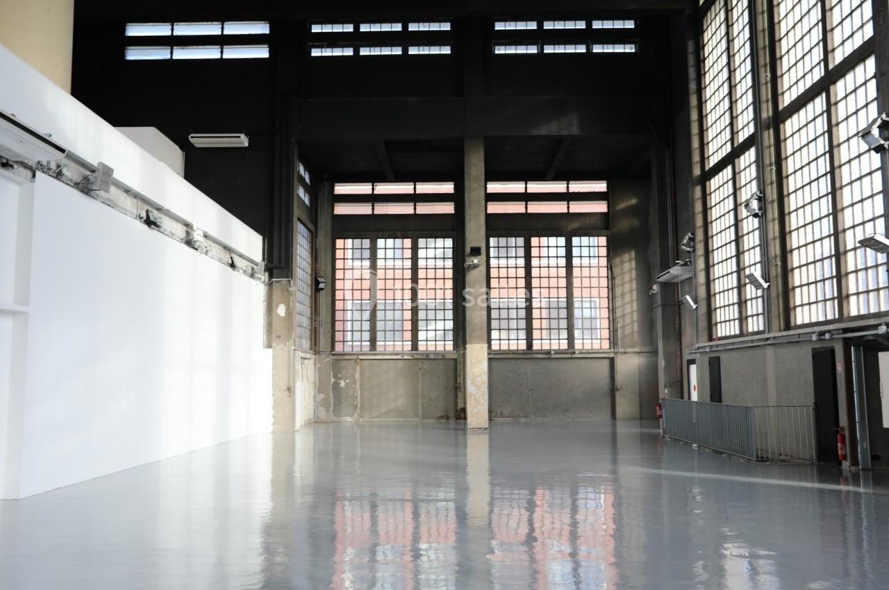 Intérieur d'un grand espace industriel avec sol brillant, murs en béton et grandes fenêtres rectangulaires.