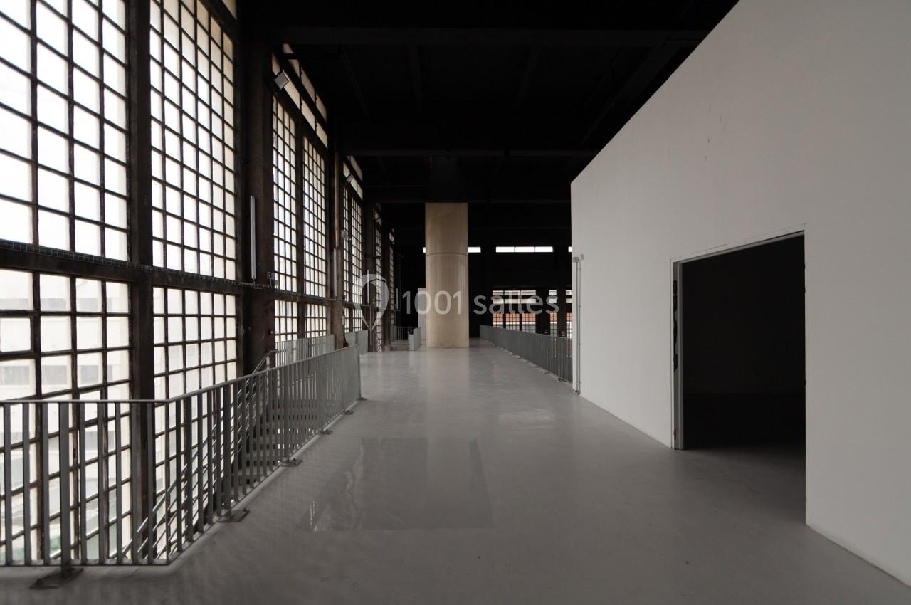 Couloir intérieur spacieux avec sol gris, murs blancs, grandes fenêtres à carreaux et garde-corps métalliques.