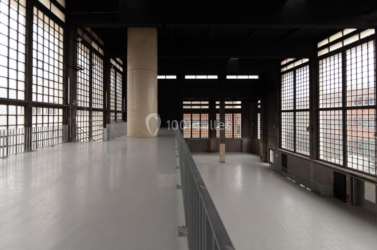 Intérieur d'un bâtiment industriel avec de grandes baies vitrées, un sol clair et des colonnes en béton.