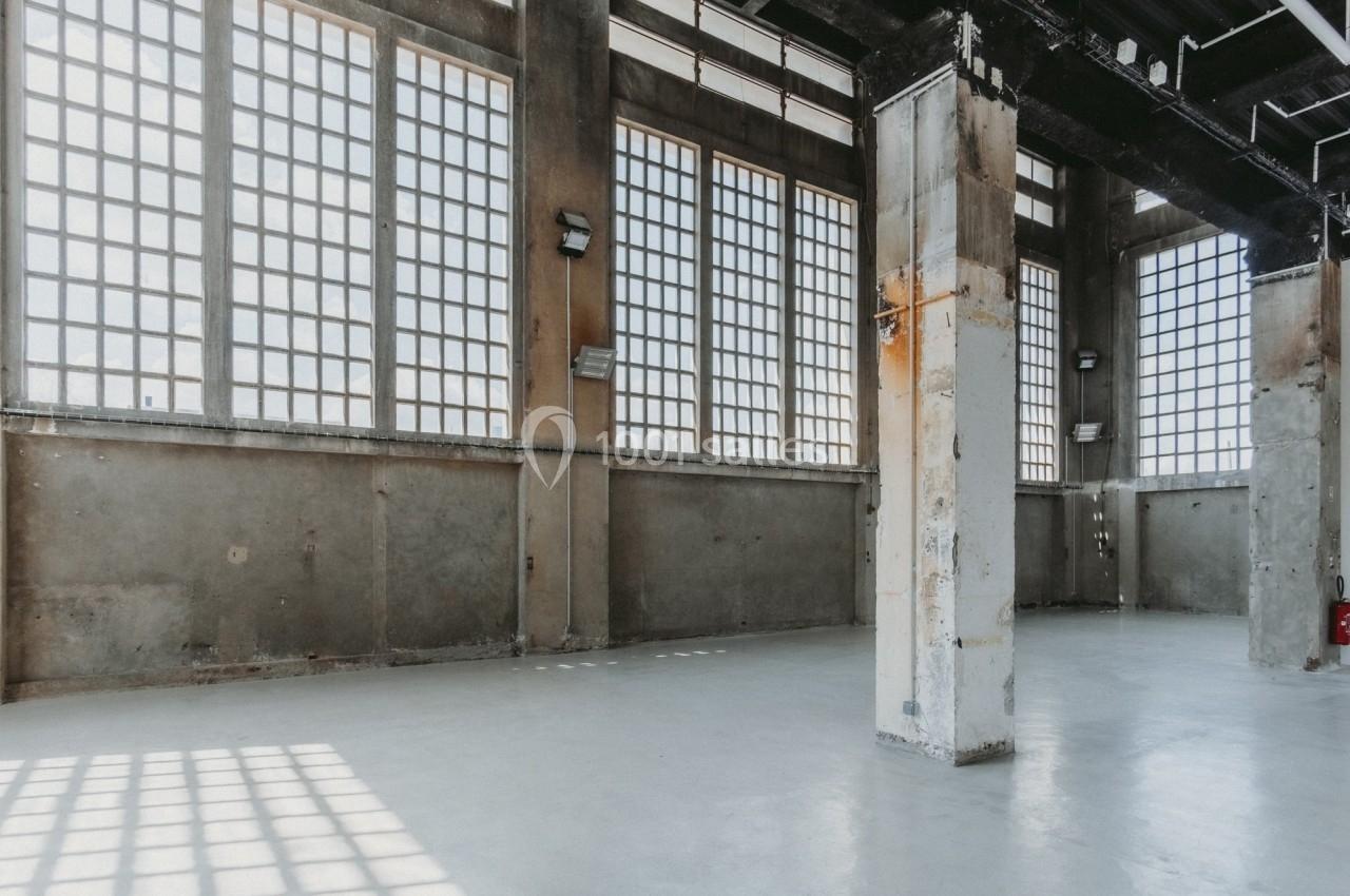 Intérieur d'un grand espace industriel vide avec des murs en béton, des colonnes et de grandes fenêtres à carreaux.