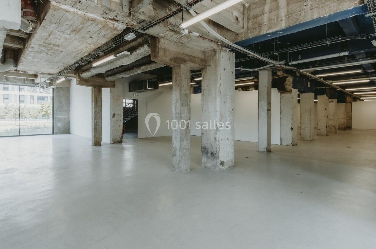 Intérieur d'un espace industriel vide avec murs en béton brut, colonnes apparentes et éclairage fluorescent.