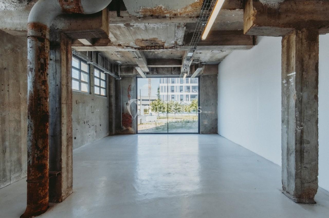 Intérieur d'un espace industriel vide avec murs en béton brut, tuyaux apparents et grande baie vitrée donnant sur l…