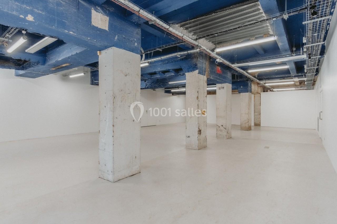 Espace intérieur vide avec colonnes en béton brut, plafond bleu et câblage apparent.
