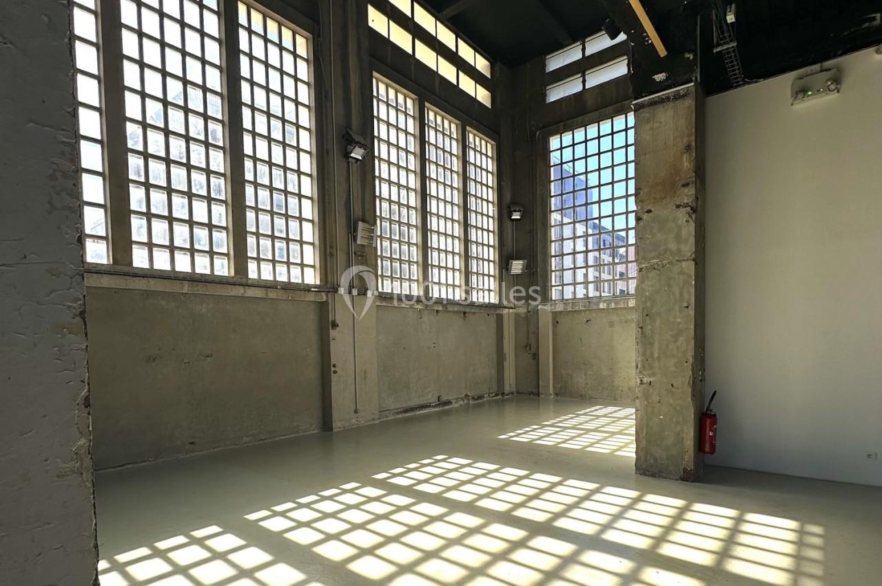 Intérieur d'une pièce lumineuse avec de grandes fenêtres à carreaux, murs en béton brut et un extincteur au fond.