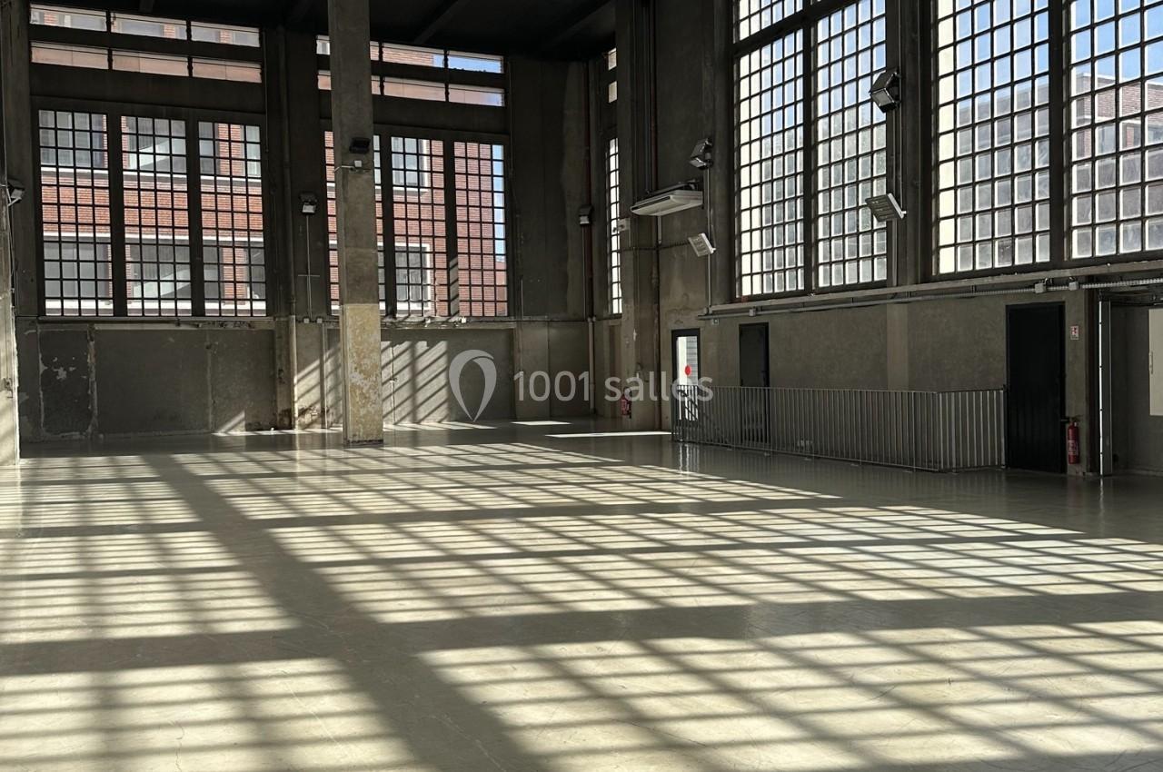 Grande salle vide avec de hauts plafonds, murs en béton et larges fenêtres laissant entrer une lumière naturelle.