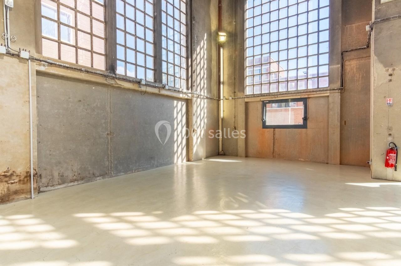 Intérieur d'un grand espace industriel vide avec murs en béton, grandes fenêtres et sol clair.
