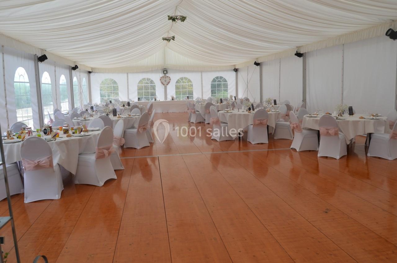 Salle de réception sous chapiteau avec tables rondes dressées, chaises blanches et sol en bois verni.