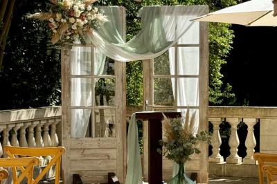 Décor de cérémonie en extérieur avec arche en bois, voilages blancs et verts, fleurs et chaises en bois.