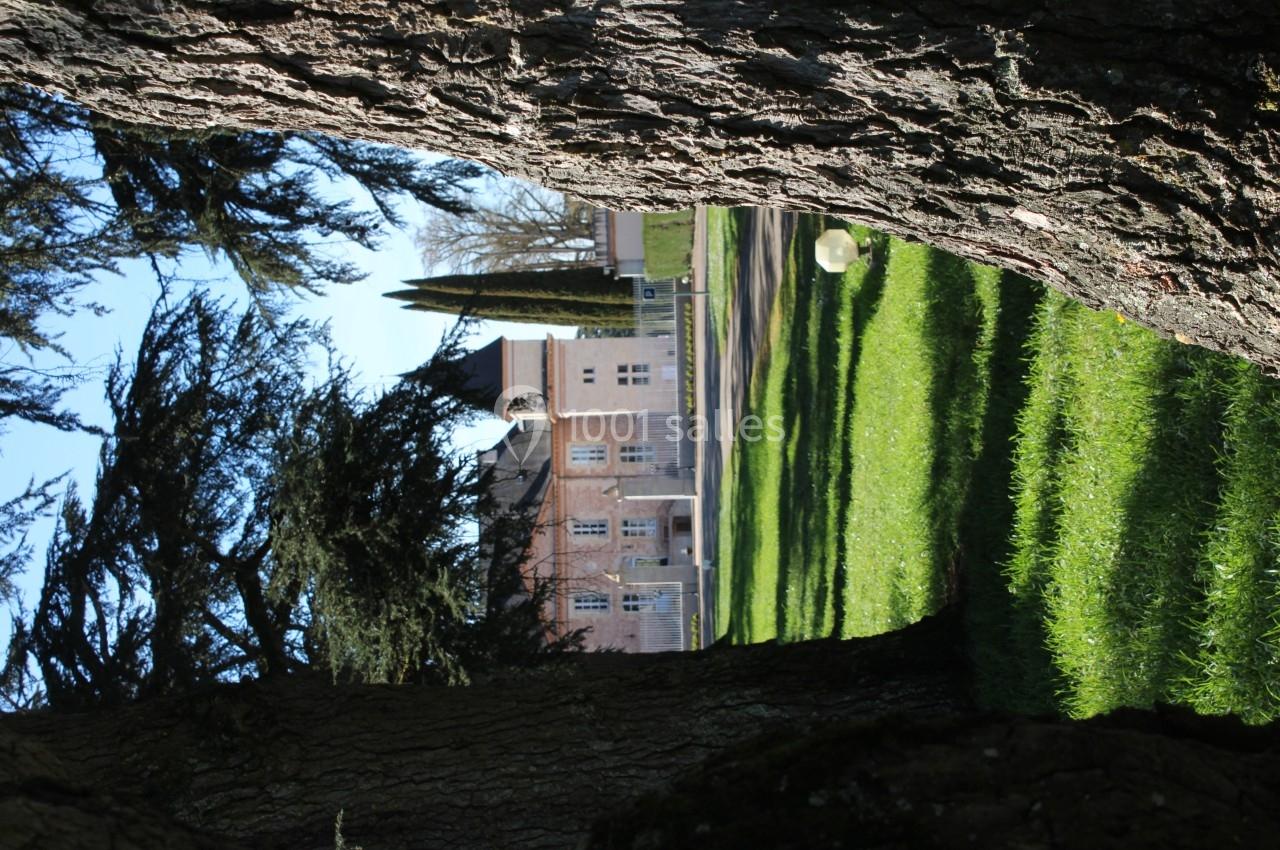 Vue d'un bâtiment en briques entouré de pelouses et d'arbres, encadré par deux troncs au premier plan.