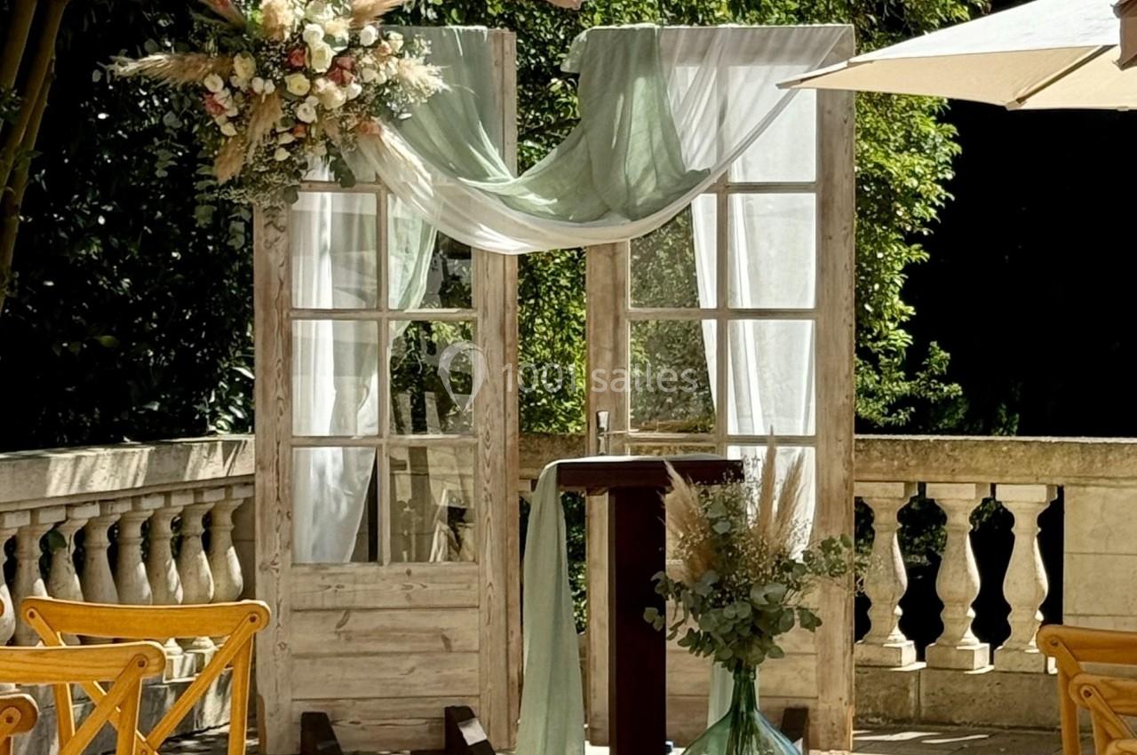 Décor de cérémonie en extérieur avec arche en bois, voilages blancs et verts, fleurs et chaises en bois.