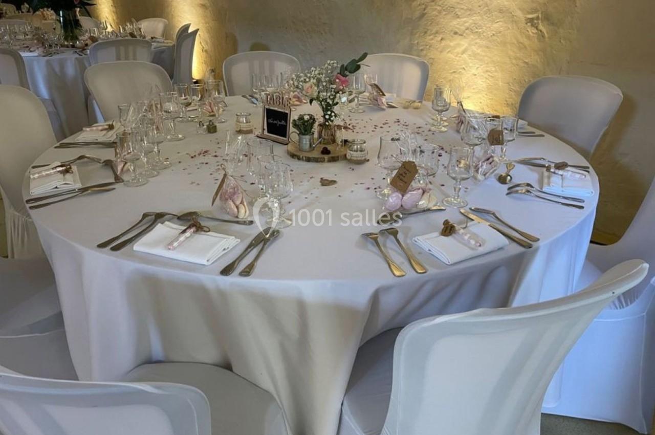 Table ronde élégamment dressée avec nappes blanches, couverts, verres et décorations florales dans une salle éclairée.
