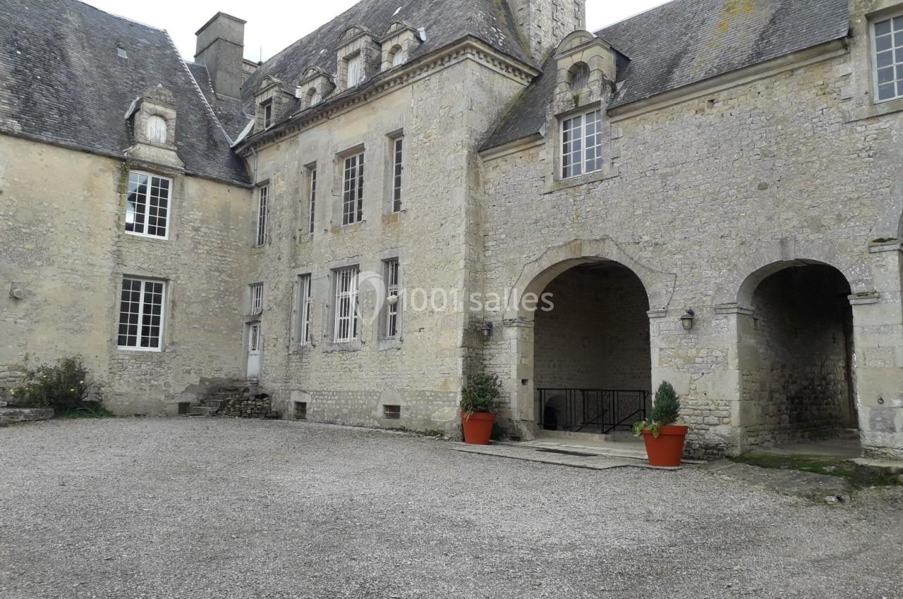 Cour intérieure d'un bâtiment en pierre de style ancien, avec des arches, des fenêtres et deux pots de fleurs rouges.