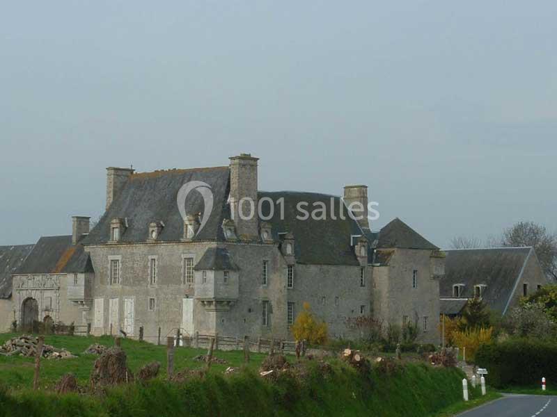 Manoir en pierre grise avec toit en ardoise, entouré de verdure et situé près d'une route de campagne.