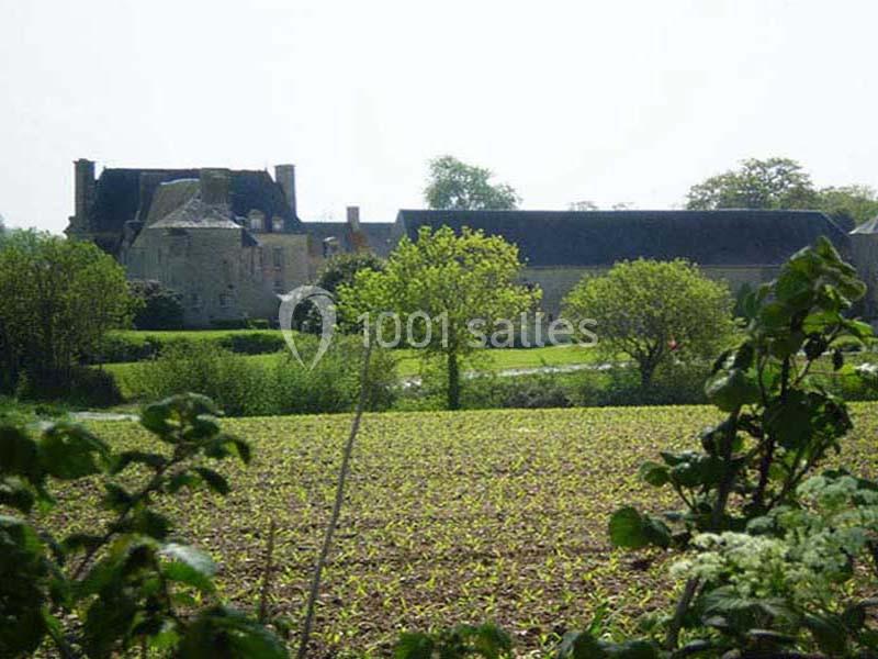 Vue d'une maison en pierre entourée de bâtiments agricoles, avec un champ cultivé et des arbres au premier plan.