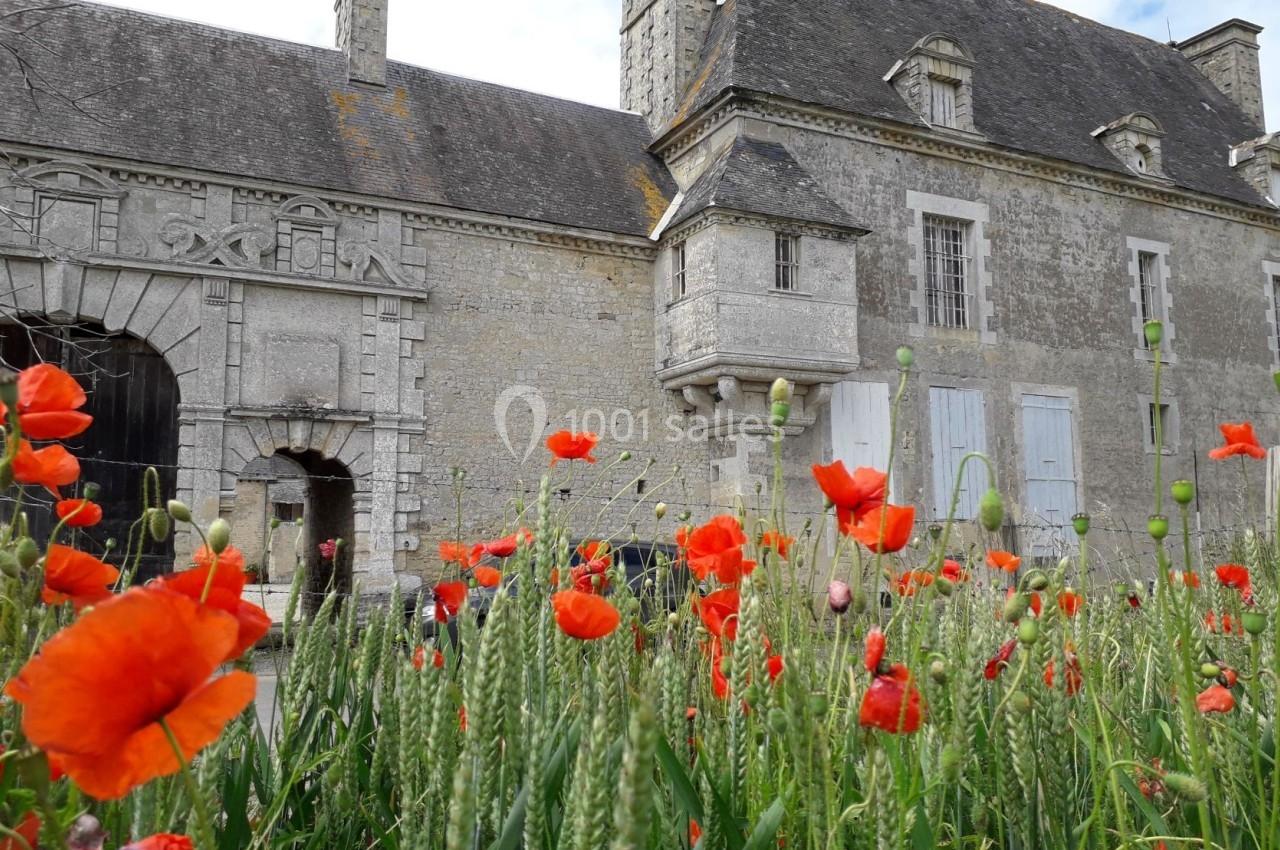 Champ de coquelicots rouges au premier plan devant un bâtiment en pierre de style ancien avec des fenêtres et une arche.